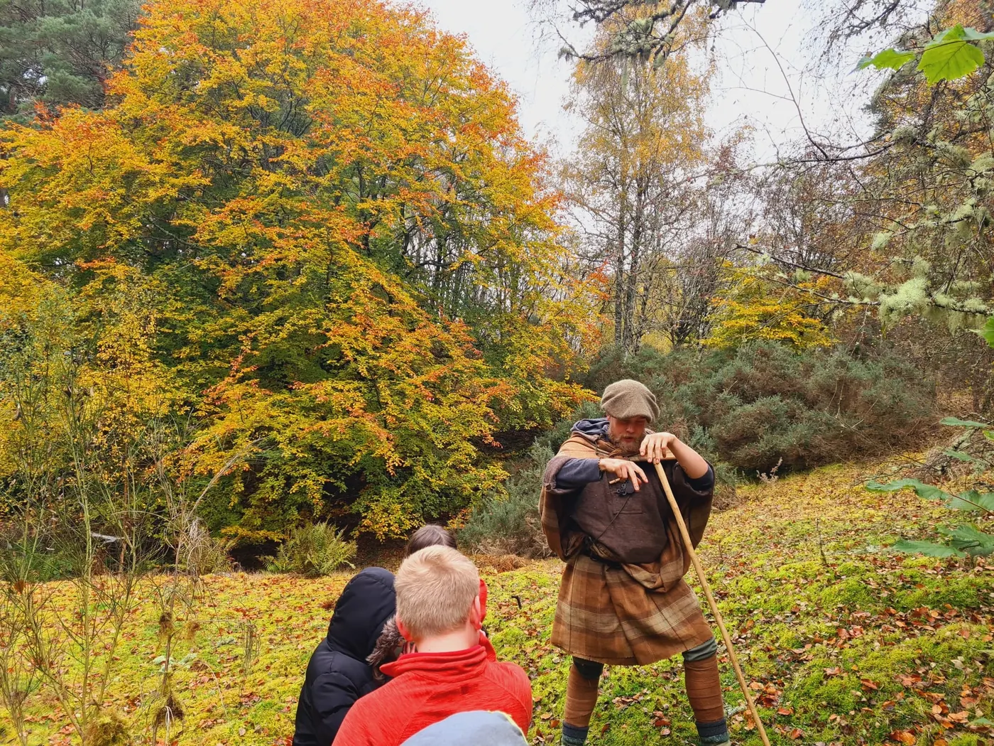 A guide in a great kilt shows a group how to work a hazel stave in autumn woodland