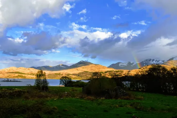 Mountains and a loch beneath a bright sky with a faint rainbow