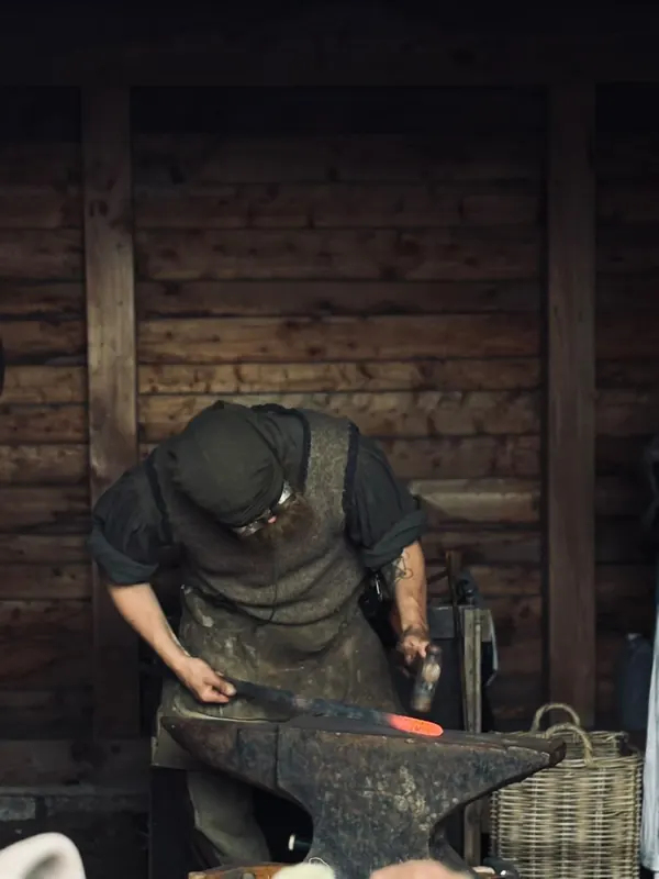 A smith working hot steel on an anvil inside a timber forge