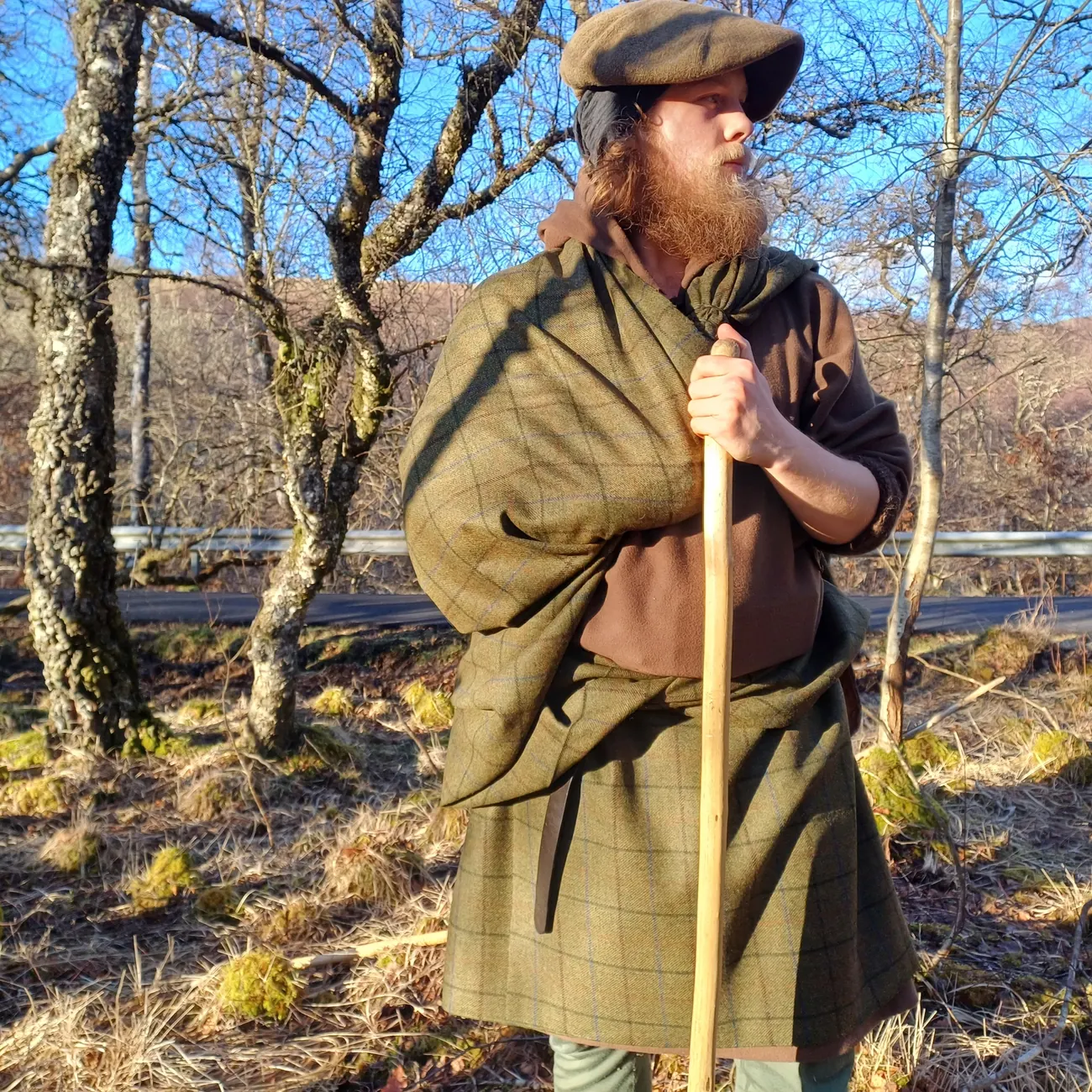 A bearded man in a belted plaid and flat cap leans on a staff among birch trees