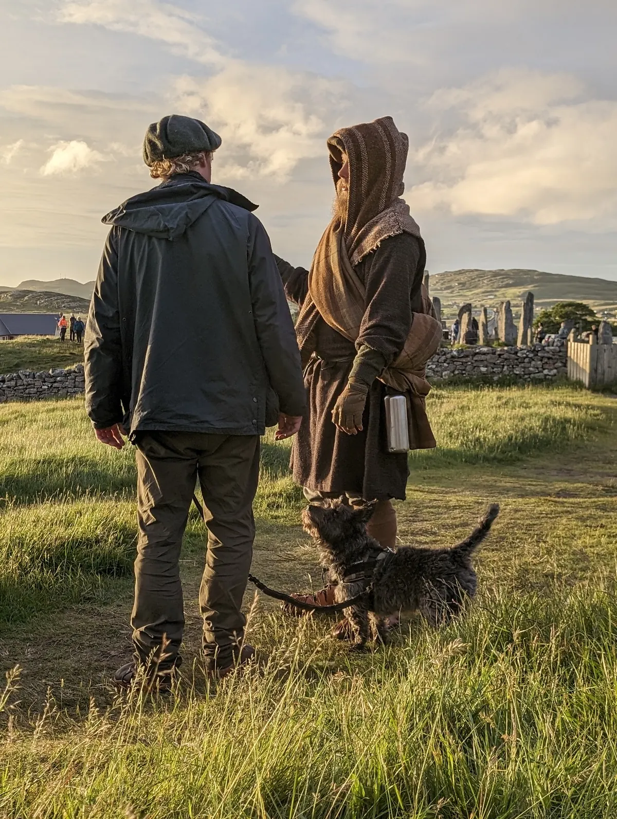 Two walkers, one in a hooded wool cloak, with a dog near a ruined chapel