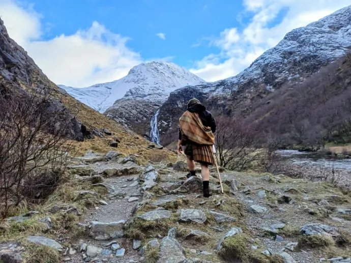 A walker in a plaid on a rocky path below snow-dusted mountains